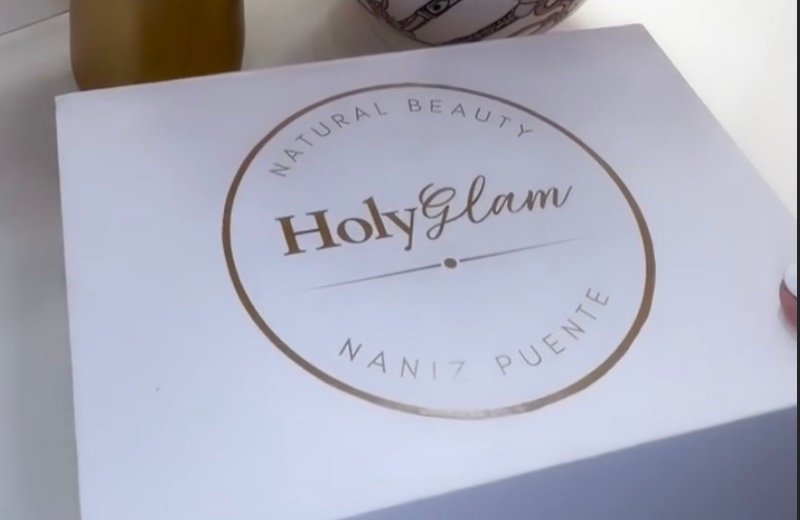 Caja blanca con el logo HolyGlam Natural Beauty, Naniz Puente, junto a un jarrón y flores.